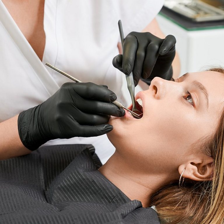 Female dentist in black sterile gloves examining patient teeth with dental explorer and mirror. Young woman lying in dental chair while having prophylactic examination. Concept of dentistry.